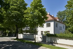 Straßenansicht der Villa hinter verputzter Einfriedungsmauer mit altem Baumbestand