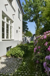 Helle Seitenfassade mit Sprossenfenstern und gepflastertem Gartenweg entlang blühender Rhododendren