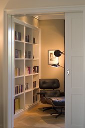 Bibliotheksnische mit Schiebetür, weißem Einbauregal, Fischgrätparkett und klassischem Loungesessel