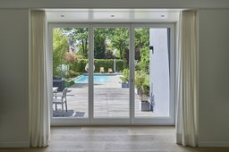 Blick aus dem Wohnzimmer durch bodentiefe Verglasung auf Holzterrasse und Pool