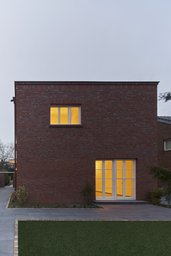 Frontale Ansicht der zweigeschossigen kubischen Klinkerfassade mit beleuchtetem Terrassenzugang im Abendlicht