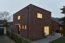 Zweigeschossiger kubischer Klinkerbau mit beleuchtetem Eingangsbereich und Fenstern in der Dämmerung