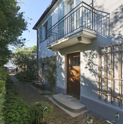 Eingangsfassade mit Eichentür, Natursteinstufe, Balkon auf Konsolen und berankte Holzspaliere