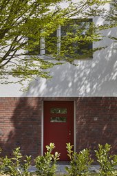 Rote Eingangstür im Backsteinsockel unter weißer Putzfassade mit Baumschatten