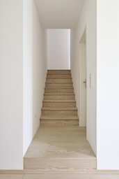 Geradlinige Holztreppe zwischen weißen Wänden zum Obergeschoss