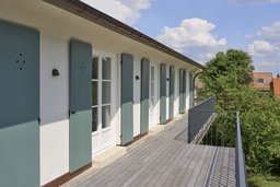 Langgestreckter Balkon im Obergeschoss mit Holzbelag, Metallgeländer und salbeifarbenen Fensterläden